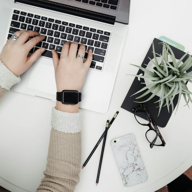 Hände tippen auf einer Laptop-Tastatur, daneben eine Pflanze, ein Handy und Stifte.